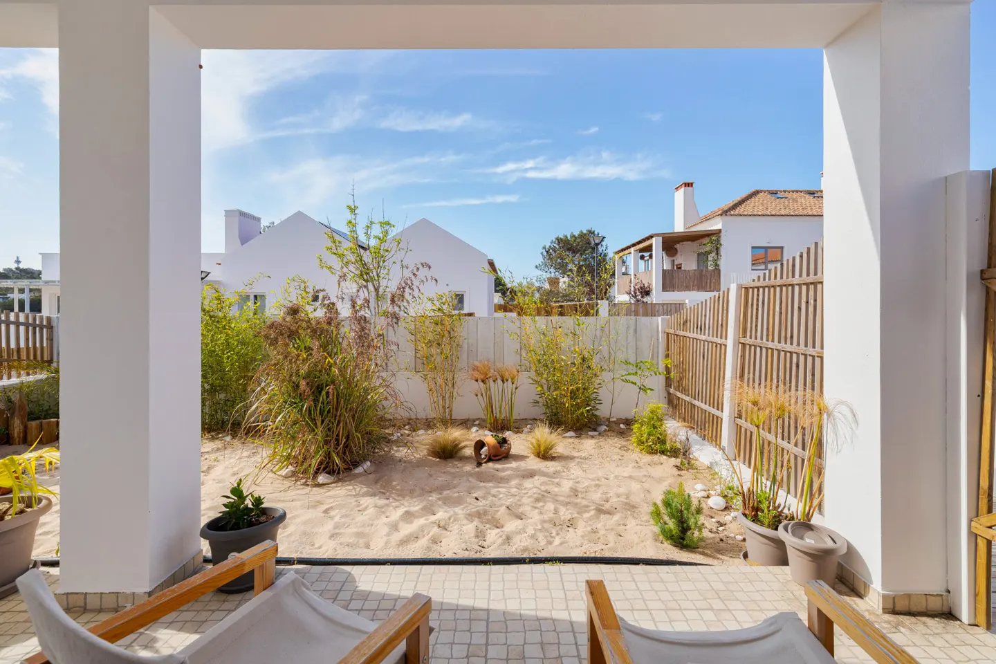 View from a patio with white columns to a sandy backyard with plants and a wooden fence. White houses are visible in the background under a blue sky.