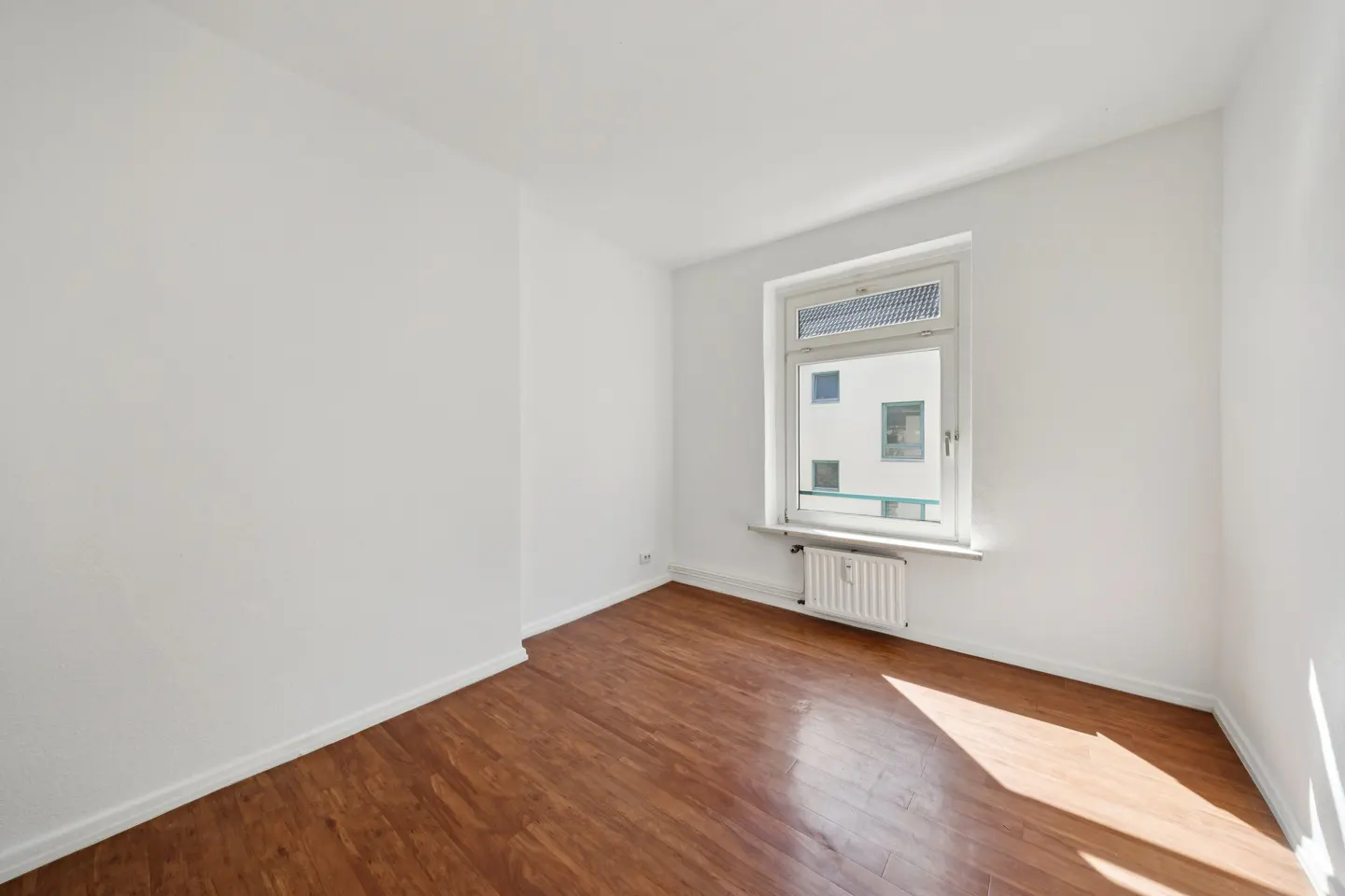 Bright, empty room with white walls, wood floors, and a window with a radiator below.