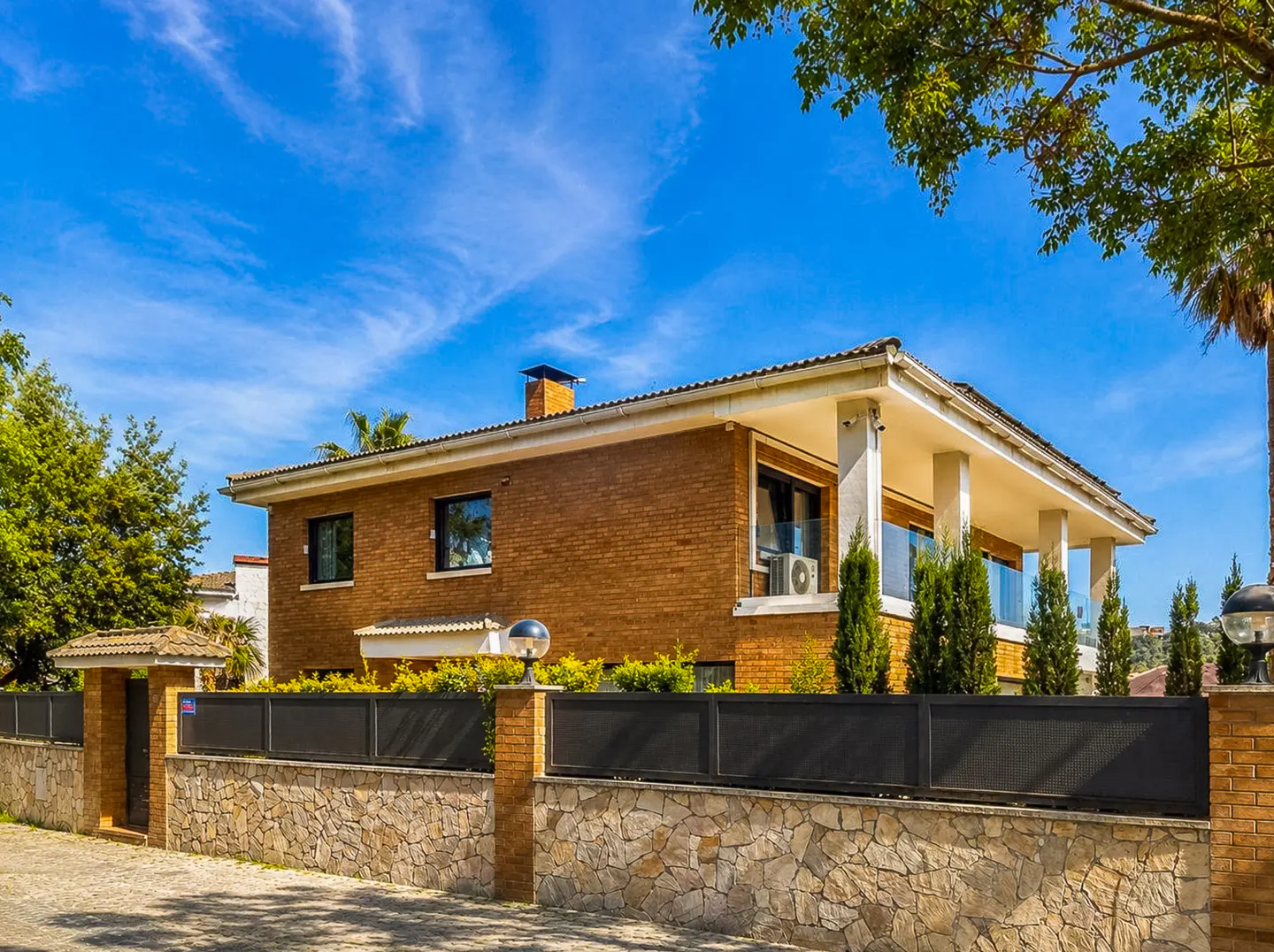 Two-story brick house with a white porch roof, stone wall, and black metal fence under a blue sky.