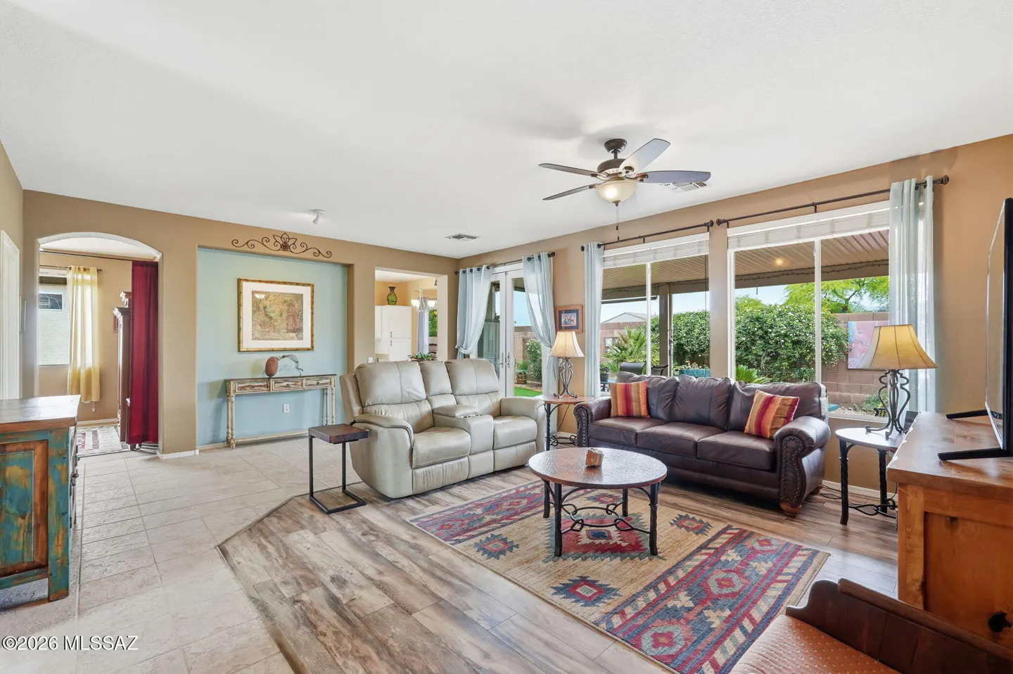 Living room with tan walls, tile and wood floors, a leather sofa and loveseat, and a patterned rug. Large windows look out onto a green backyard.