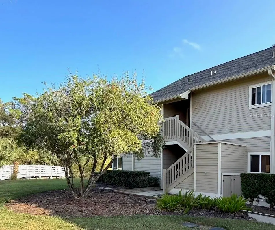 Tan two-story condo with gray roof and wooden stairs leading to the second floor. A tree is in the front yard.