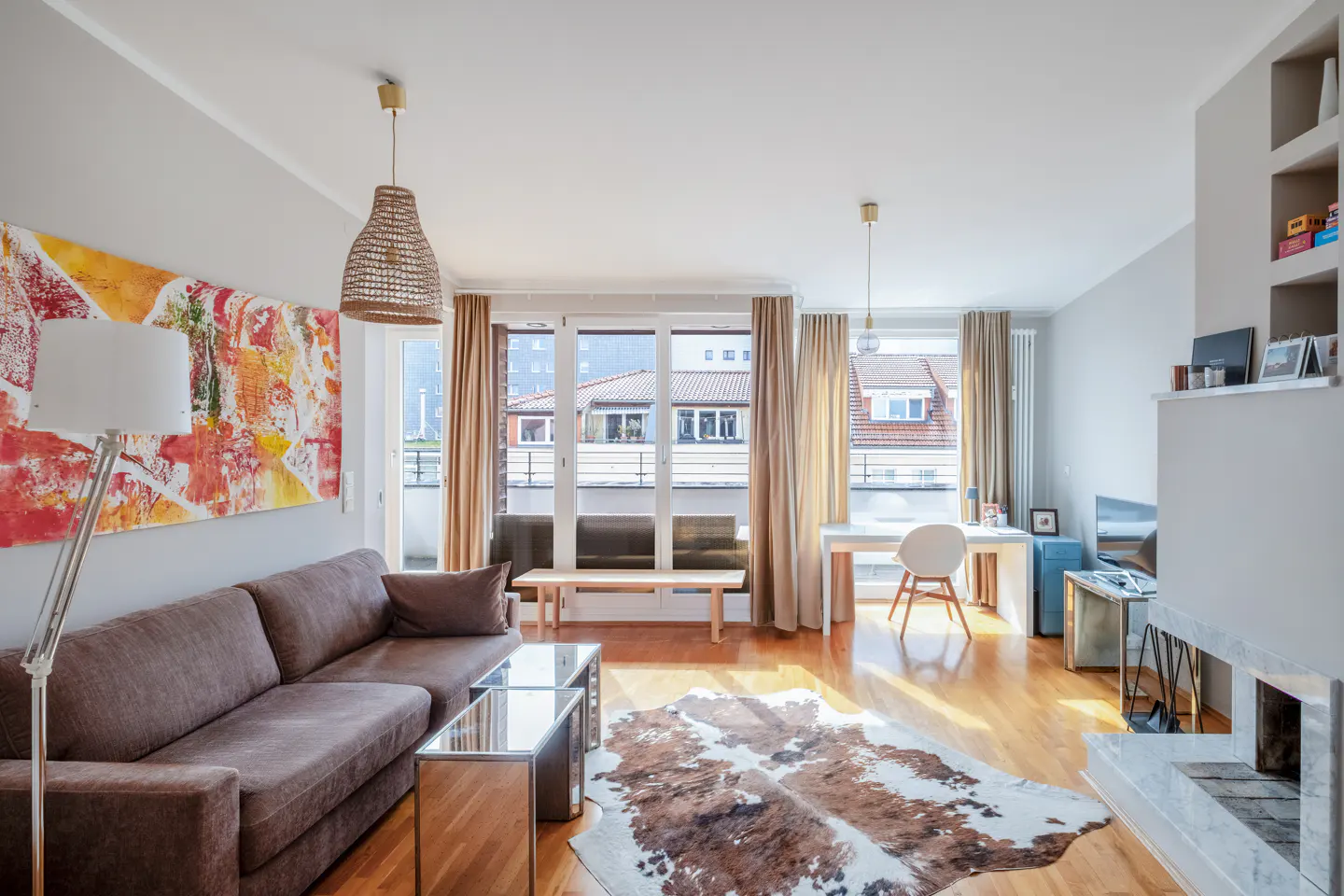 Bright living room with a brown sofa, a cowhide rug, and a modern fireplace. Large windows with beige curtains let in natural light.
