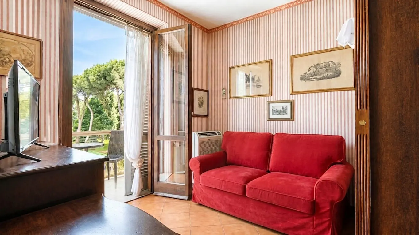 A cozy room with a red sofa, striped wallpaper, framed art, and a glass door opening to a sunny balcony with trees.