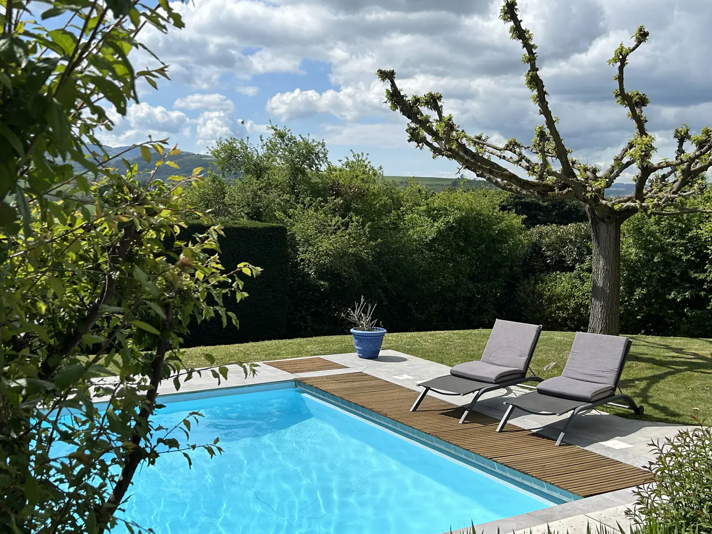 Outdoor pool with turquoise water, two gray lounge chairs, wood decking, green lawn, trees, and cloudy sky.