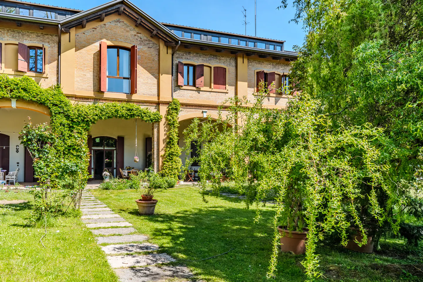 Exterior of a two-story tan brick house with brown shutters, green lawn, and stone walkway.