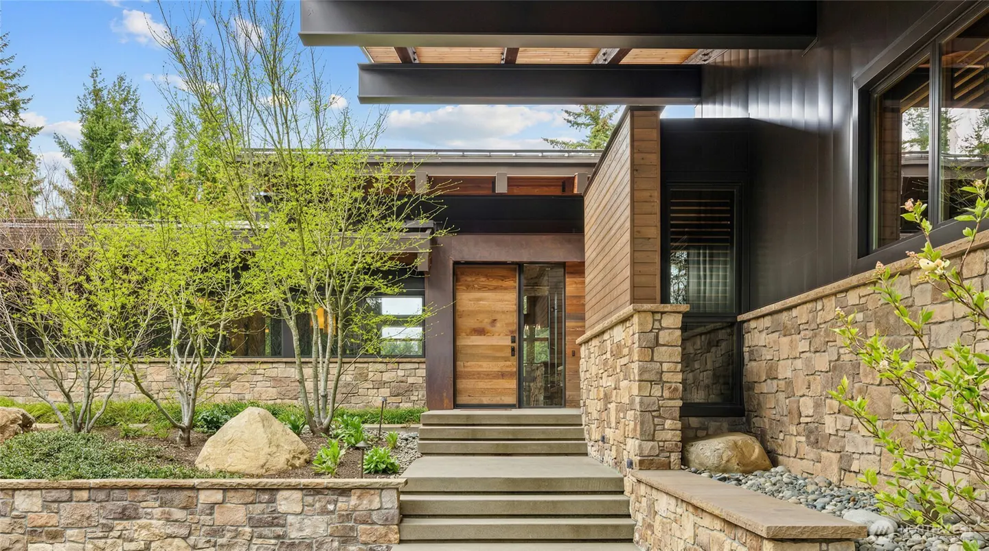 Modern home exterior with stone accents, a wooden door, and concrete steps leading to the entrance. Trees and greenery surround the house.