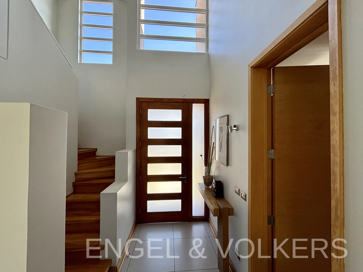 Bright foyer with white walls, wood stairs, and a modern wood door with glass panels. A small table sits against the wall with decor.