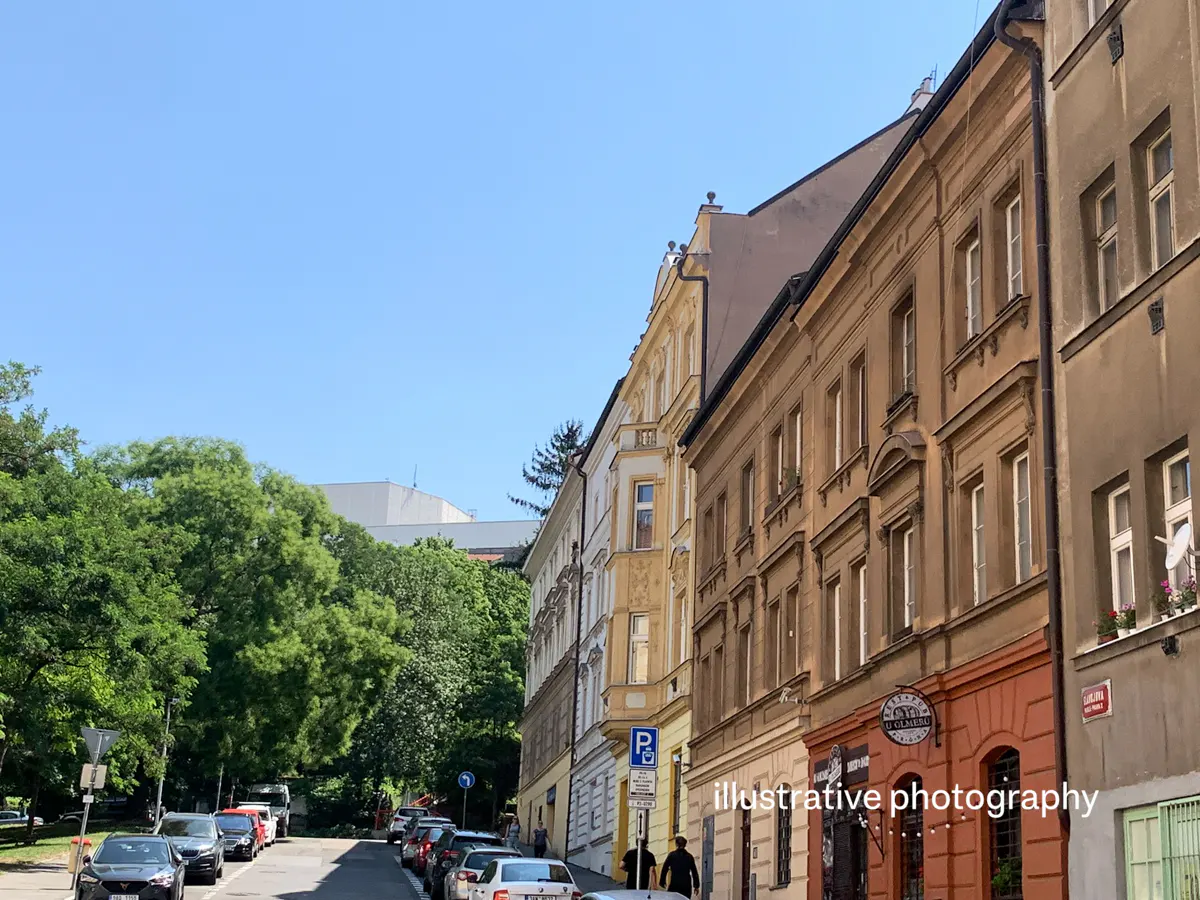 Street view of Prague with parked cars, trees, and buildings of various colors under a clear blue sky.