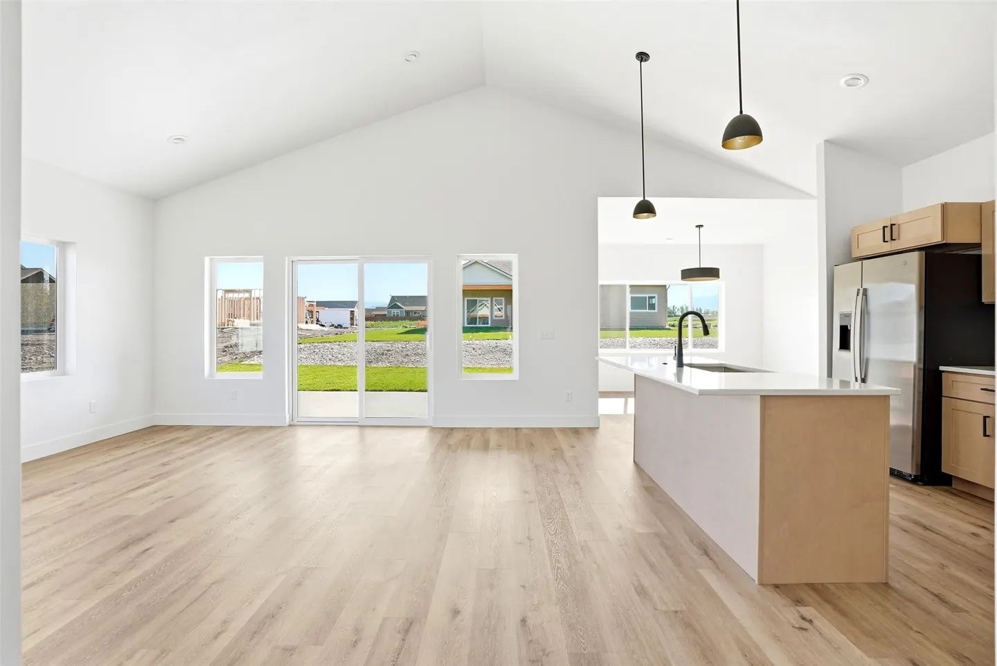 Bright, open-concept home interior with white walls, light wood floors, and a kitchen island. Windows offer views of the green lawn and neighborhood.