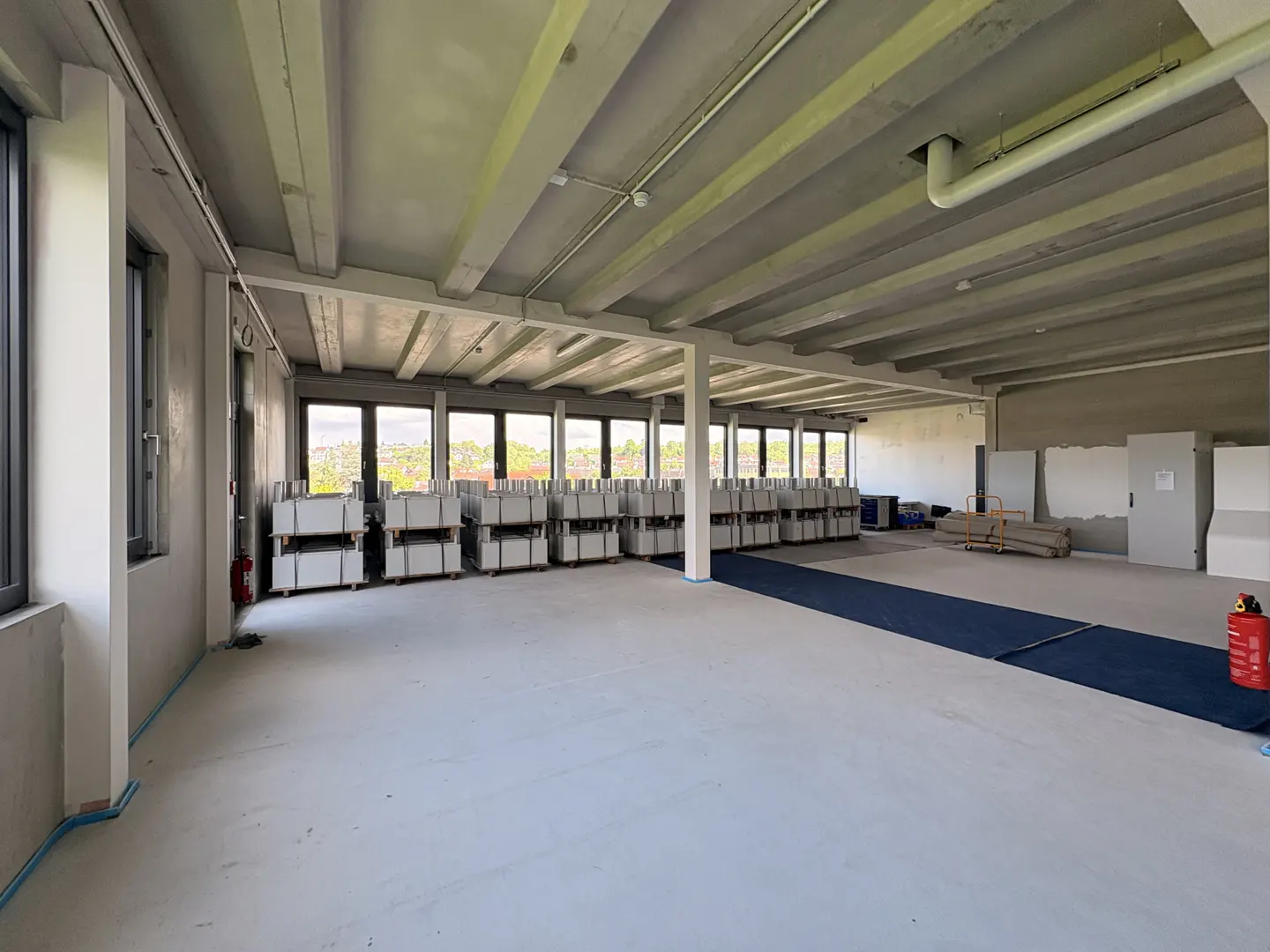 A large, empty room with gray concrete floors and ceiling, stacks of white boxes, and a blue carpet.
