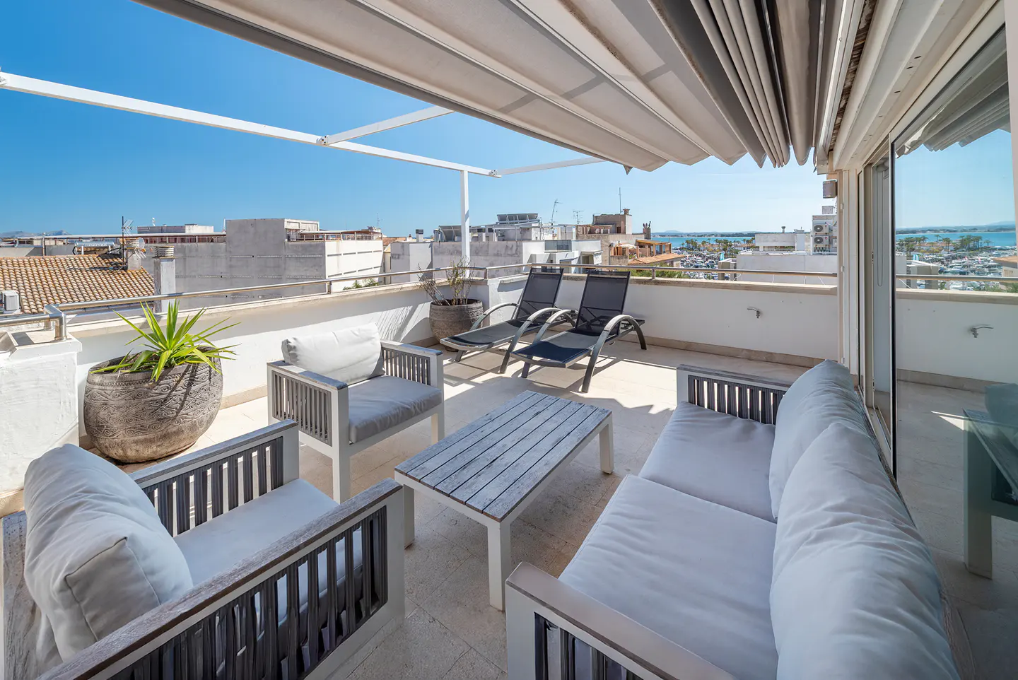 A rooftop patio with white furniture, including a sofa, chairs, and a table, under a retractable awning, with a city and ocean view.
