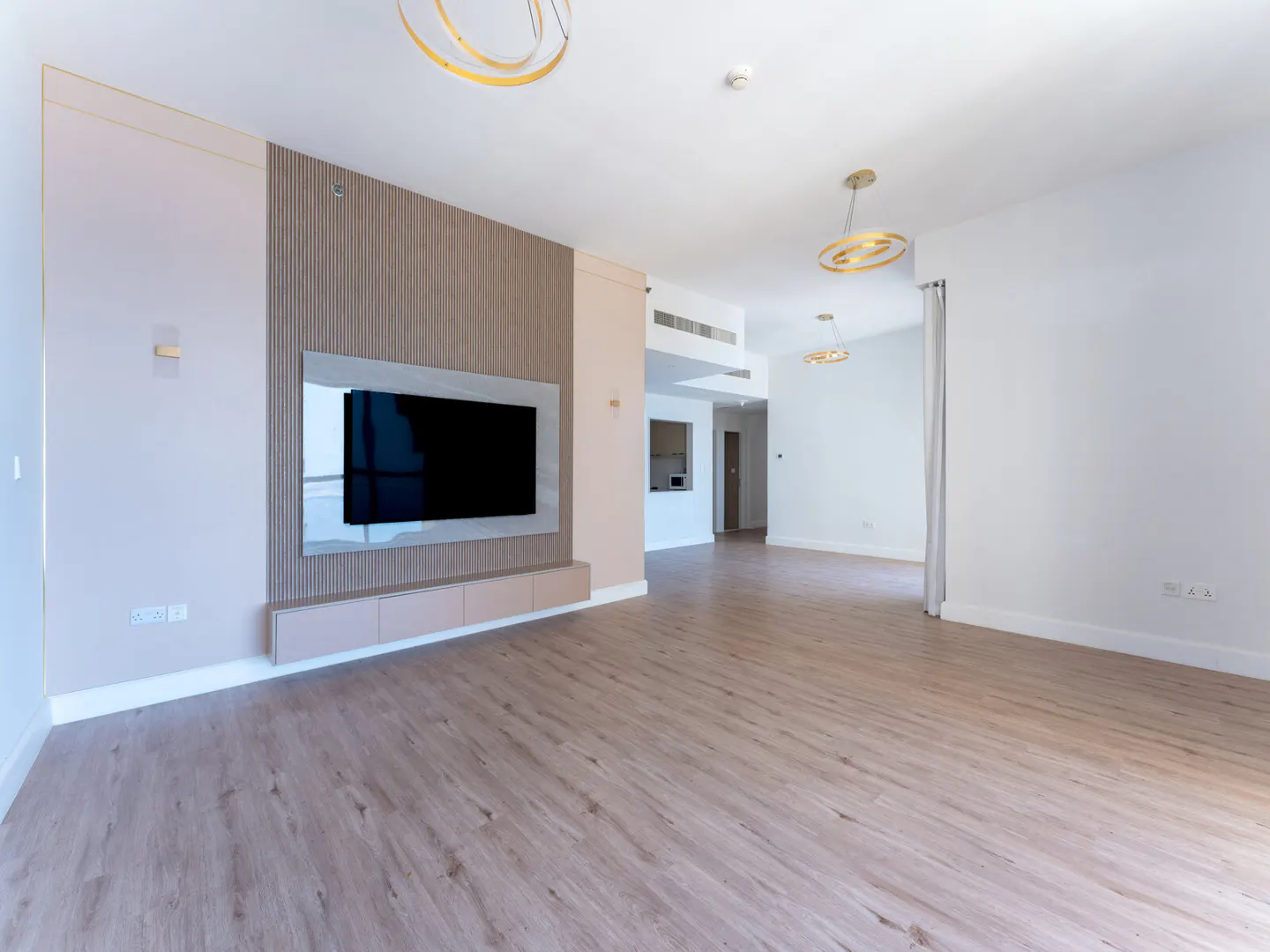A bright, empty living room with wood floors, a large TV on the wall, and modern gold light fixtures.