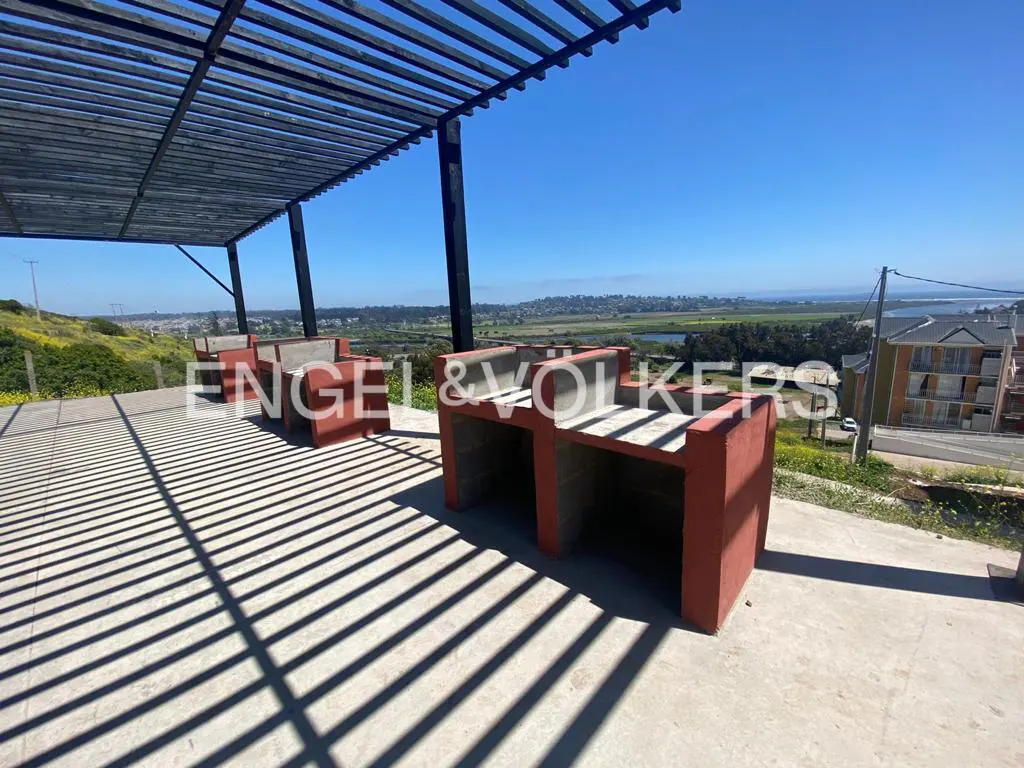 Outdoor patio with red concrete benches and a pergola casting shadows on the ground, overlooking a green landscape and blue sky.