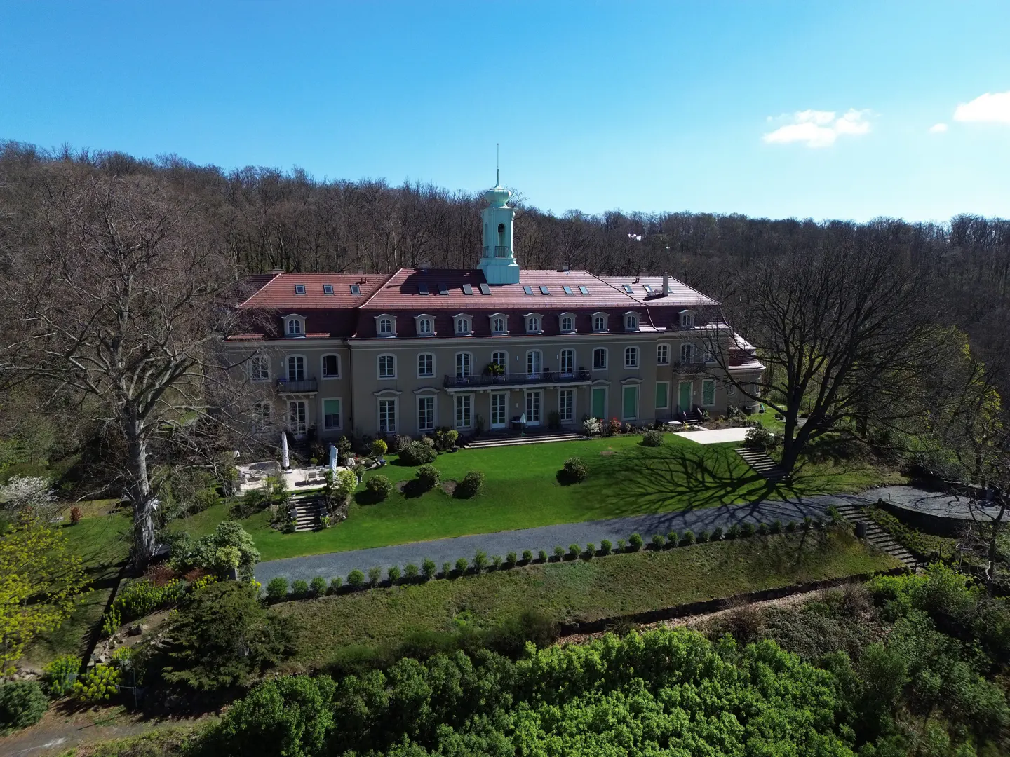 Aerial view of a large, beige building with a red roof and a green-topped tower, surrounded by green lawns and trees.