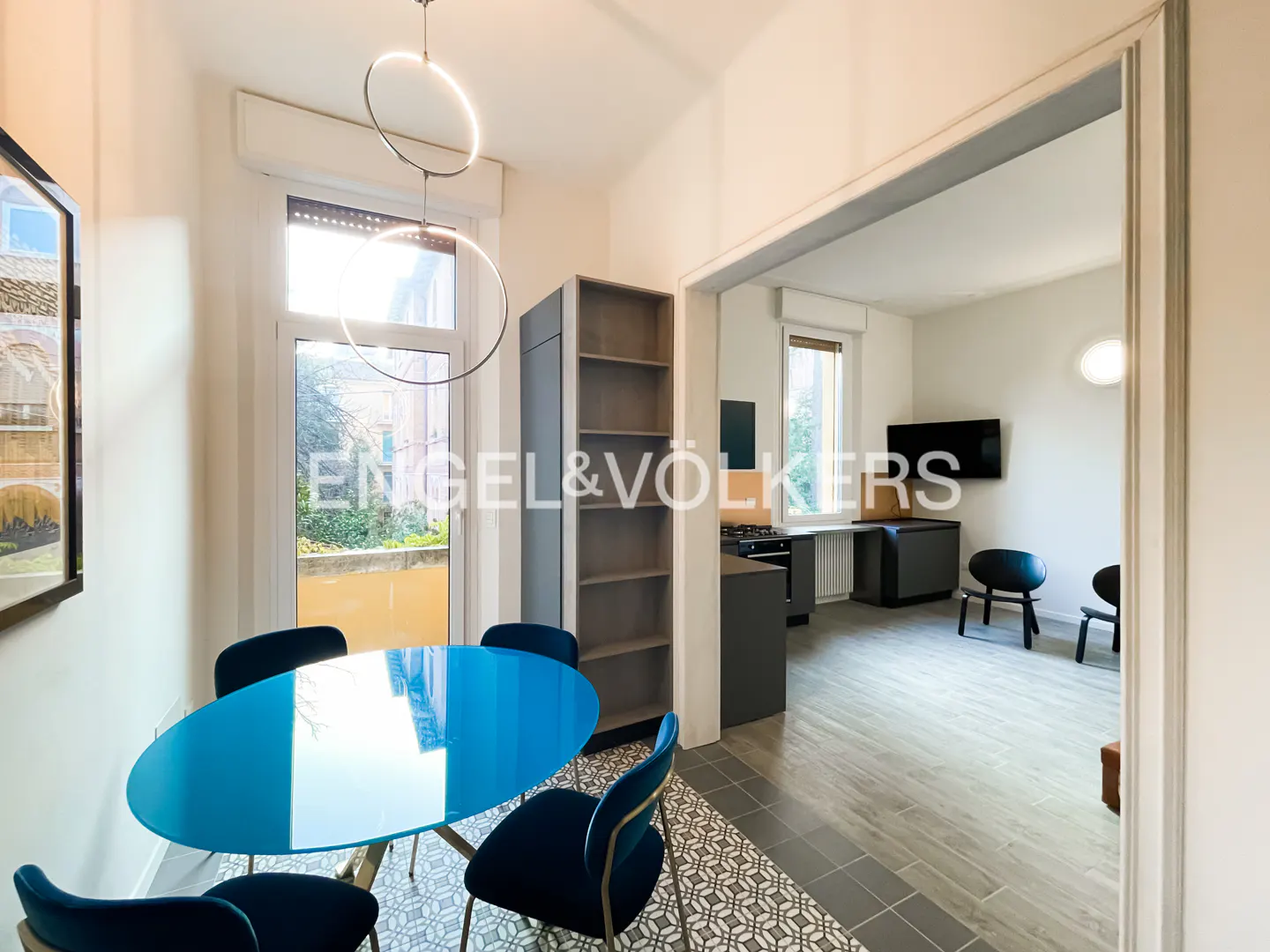 A bright, modern dining room with a blue oval table, navy chairs, and a gray bookshelf. A doorway leads to a kitchen and living area.