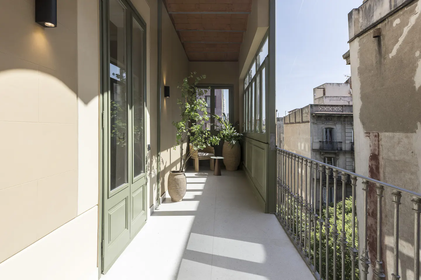 A long, narrow balcony with green trim, potted plants, and a wrought iron railing overlooks buildings.