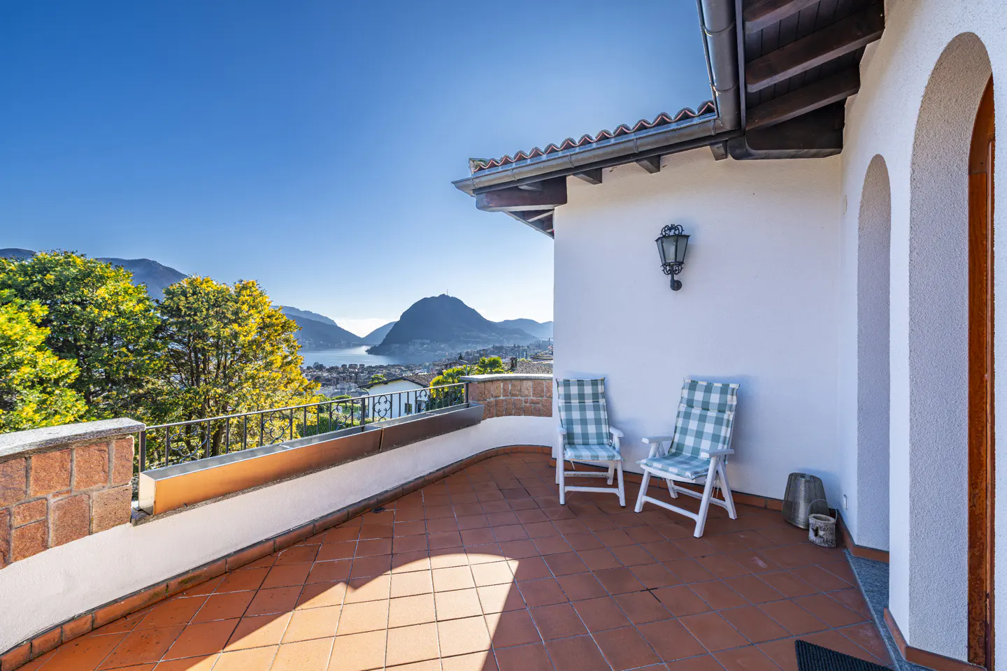 Terracotta-tiled balcony with two blue-and-white checkered chairs overlooking a lake, mountains, and trees under a clear blue sky.