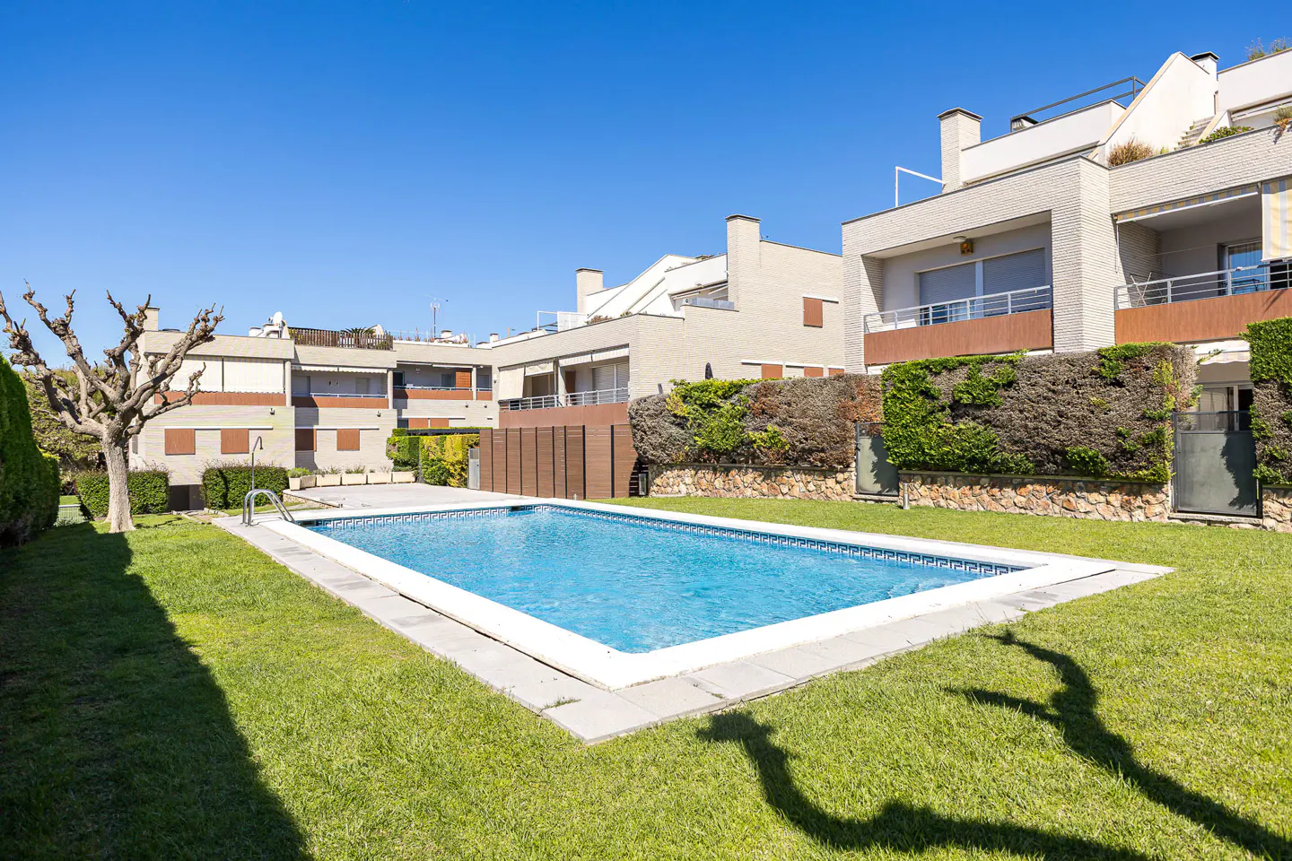 A rectangular blue swimming pool sits on a green lawn, surrounded by modern beige townhouses under a clear blue sky.