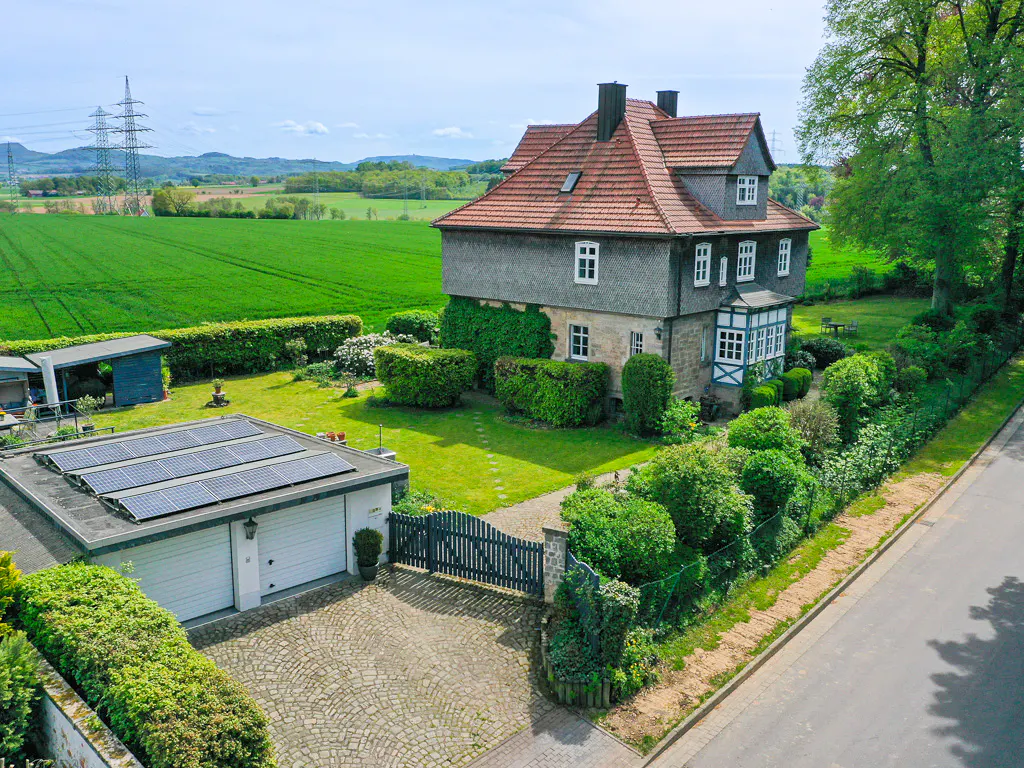 Aerial view of a two-story house with a red tile roof, solar panels on the garage, and a large green field in the background.