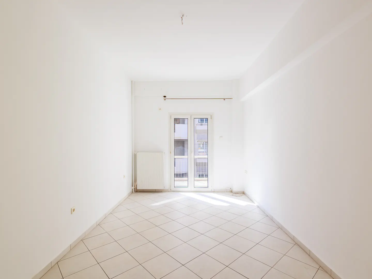 Bright, empty room with white walls, tiled floor, radiator, and balcony doors letting in sunlight.