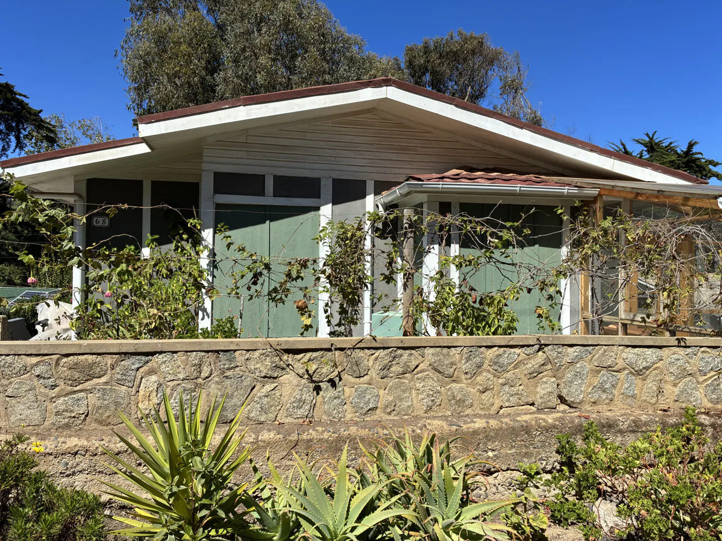 A single-story house with green shutters and a brown roof, behind a stone wall with plants.