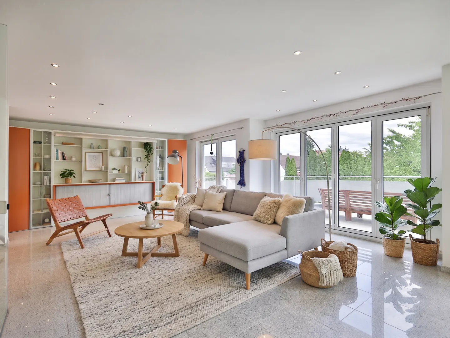 Bright living room with a gray sectional sofa, wood chair, and a round coffee table on a textured rug. Large windows overlook a balcony.