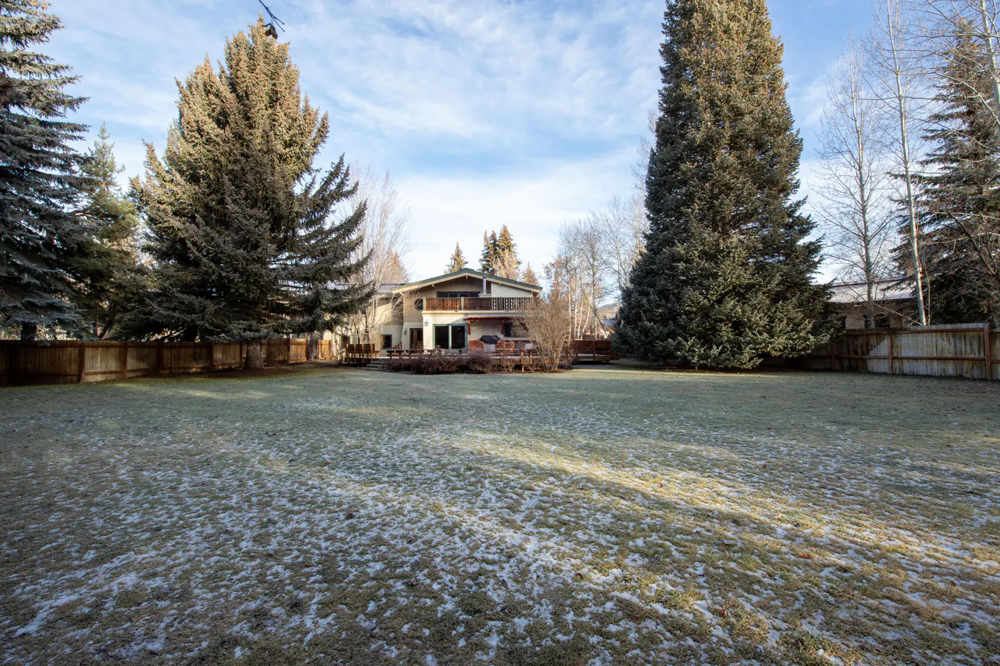 A two-story beige house with a balcony, surrounded by tall trees and a large lawn with light snow.