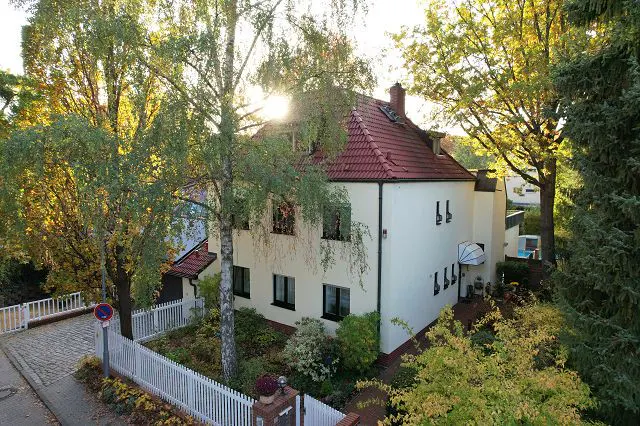 Two-story white house with a red tile roof, surrounded by trees and a white picket fence.