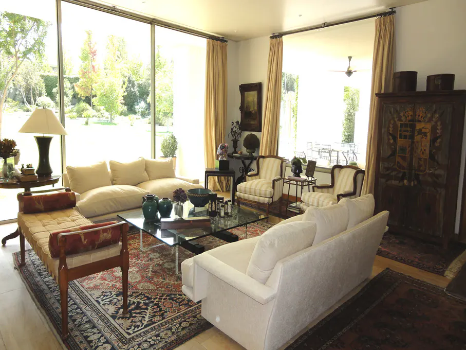 Bright living room with beige sofas, striped chairs, and a glass coffee table on an ornate rug. Large windows with tan curtains overlook a green lawn.