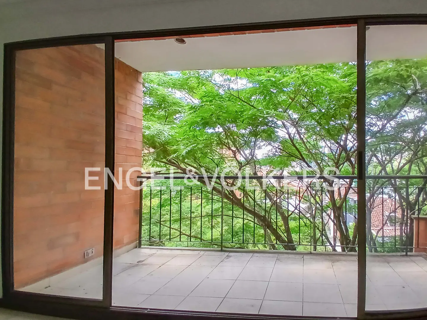Balcony view with sliding glass doors open to lush green trees and black metal railing. Tiled floor and brick wall visible.