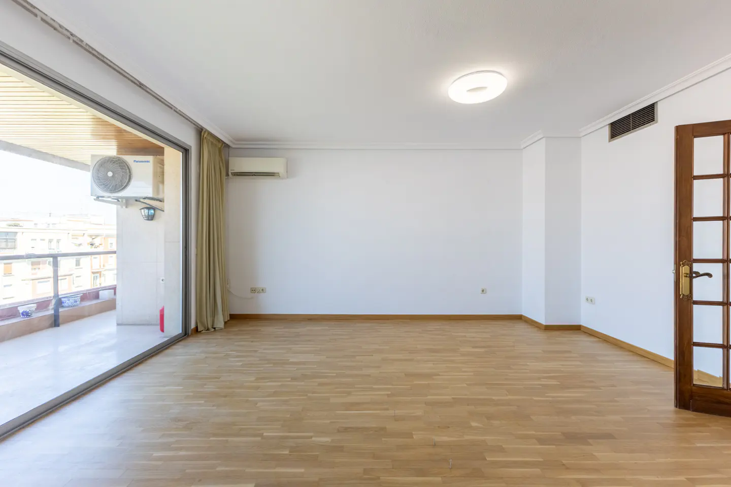 Bright, empty room with wood floors, white walls, and a sliding glass door to a balcony. A brown door is open on the right.