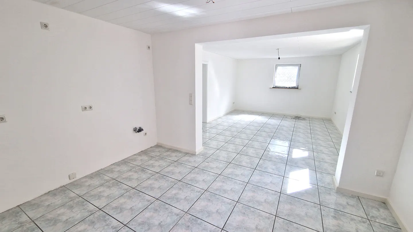 Bright, empty room with white walls and gray tiled floor. Another room is visible through a doorway.