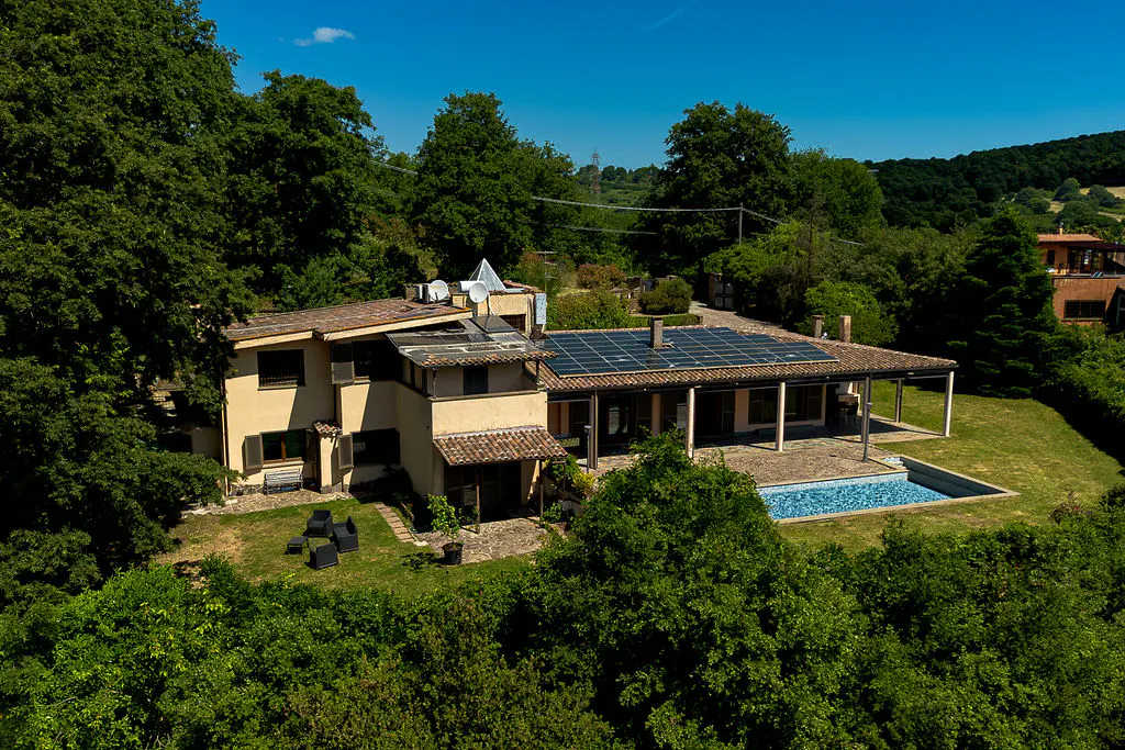 Aerial view of a beige villa with solar panels, a swimming pool, and patio furniture, surrounded by lush green trees.