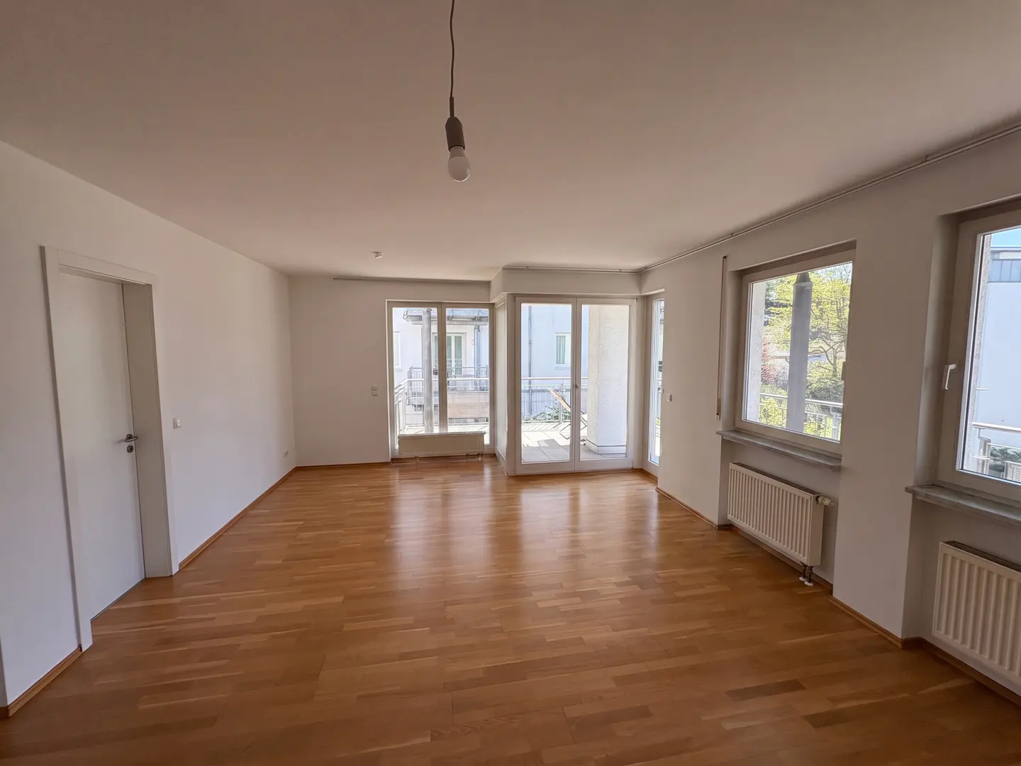 Bright, empty room with wood floors, white walls, windows, and balcony doors. A bare bulb hangs from the ceiling.
