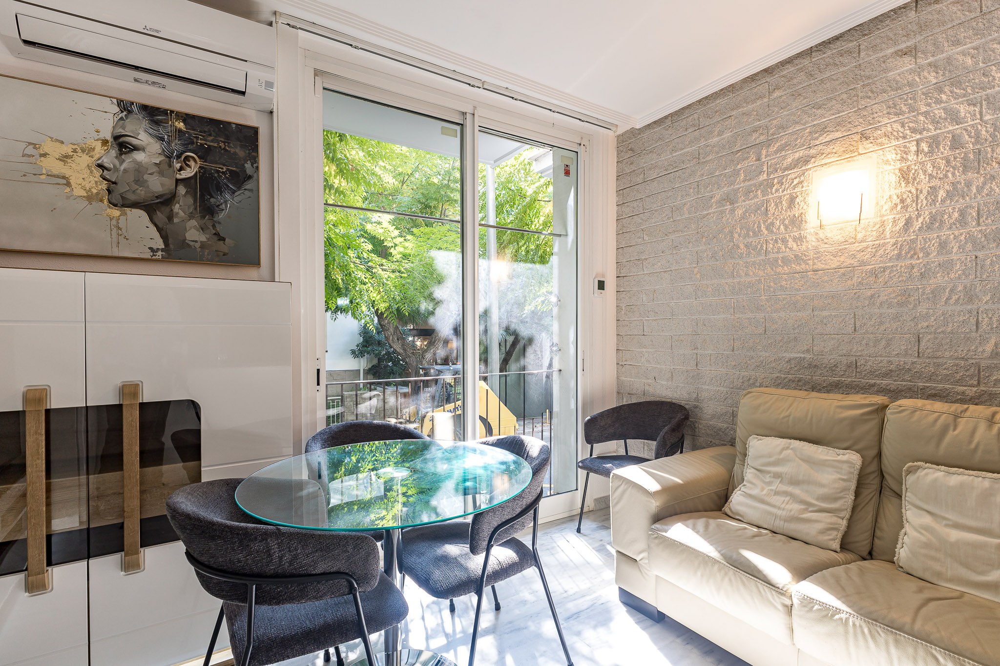 Modern living room with a beige sofa, glass dining table, dark chairs, white brick wall, and a view of trees outside. [AI-generated text]