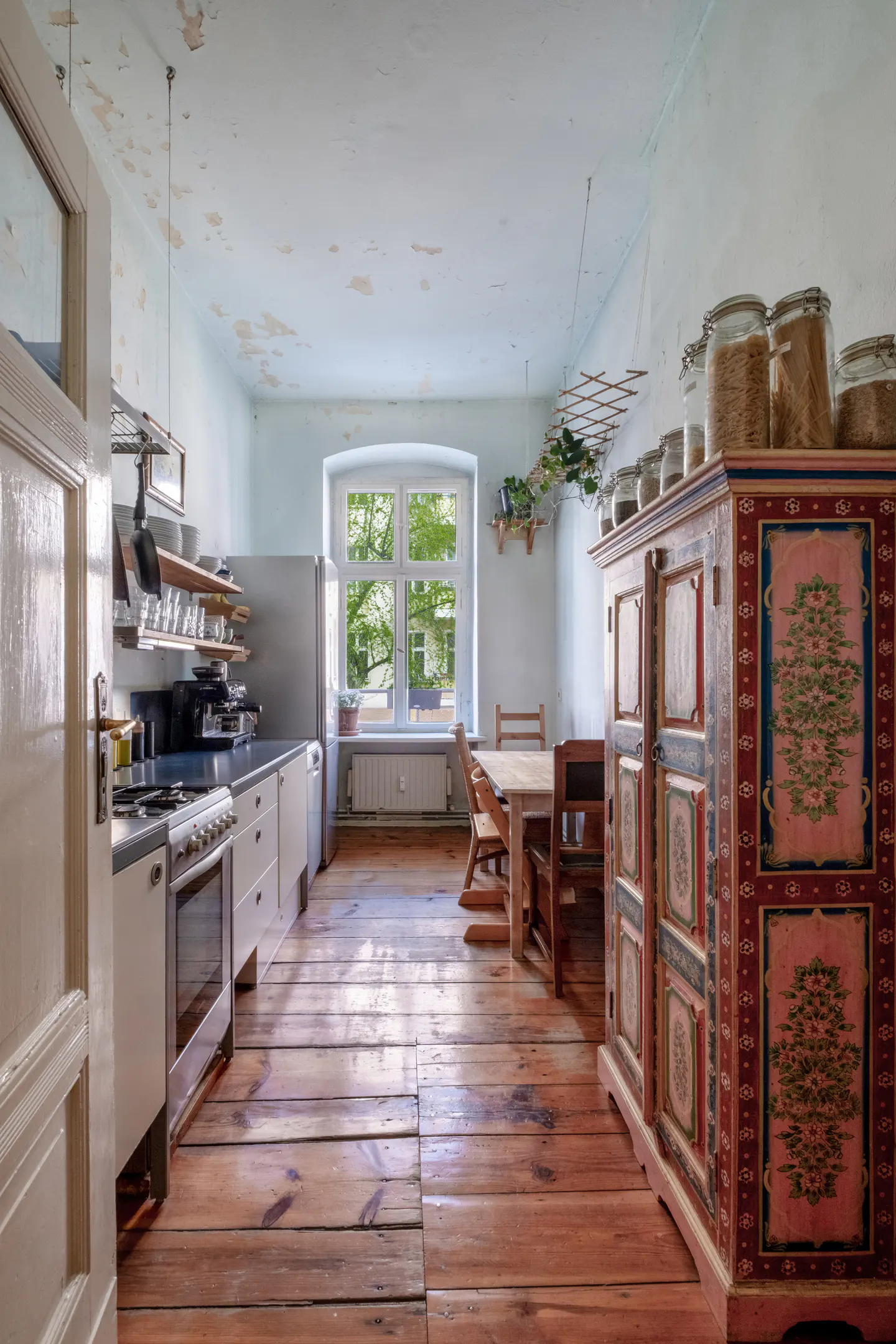 A kitchen with wood floors, white cabinets, and a painted armoire. A window overlooks a green yard.
