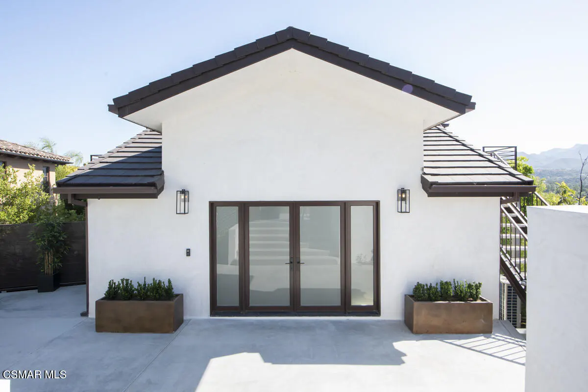 White exterior of a modern home with dark tile roof, glass double doors, and two large rectangular planters on a patio.