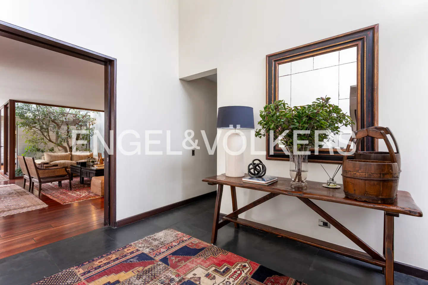 Entryway with dark wood floors, a colorful rug, and a wooden table with a lamp, vase, and wooden bucket. A living room is visible through a doorway.