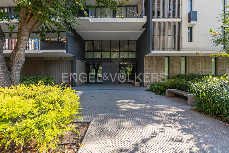 Exterior view of a modern apartment building with a gray facade and glass entrance. Green bushes and trees surround the building.