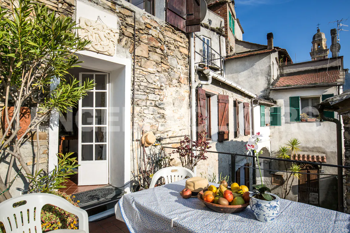 Outdoor patio with stone wall, white door, and table with fruit bowl. Buildings and blue sky in the background.