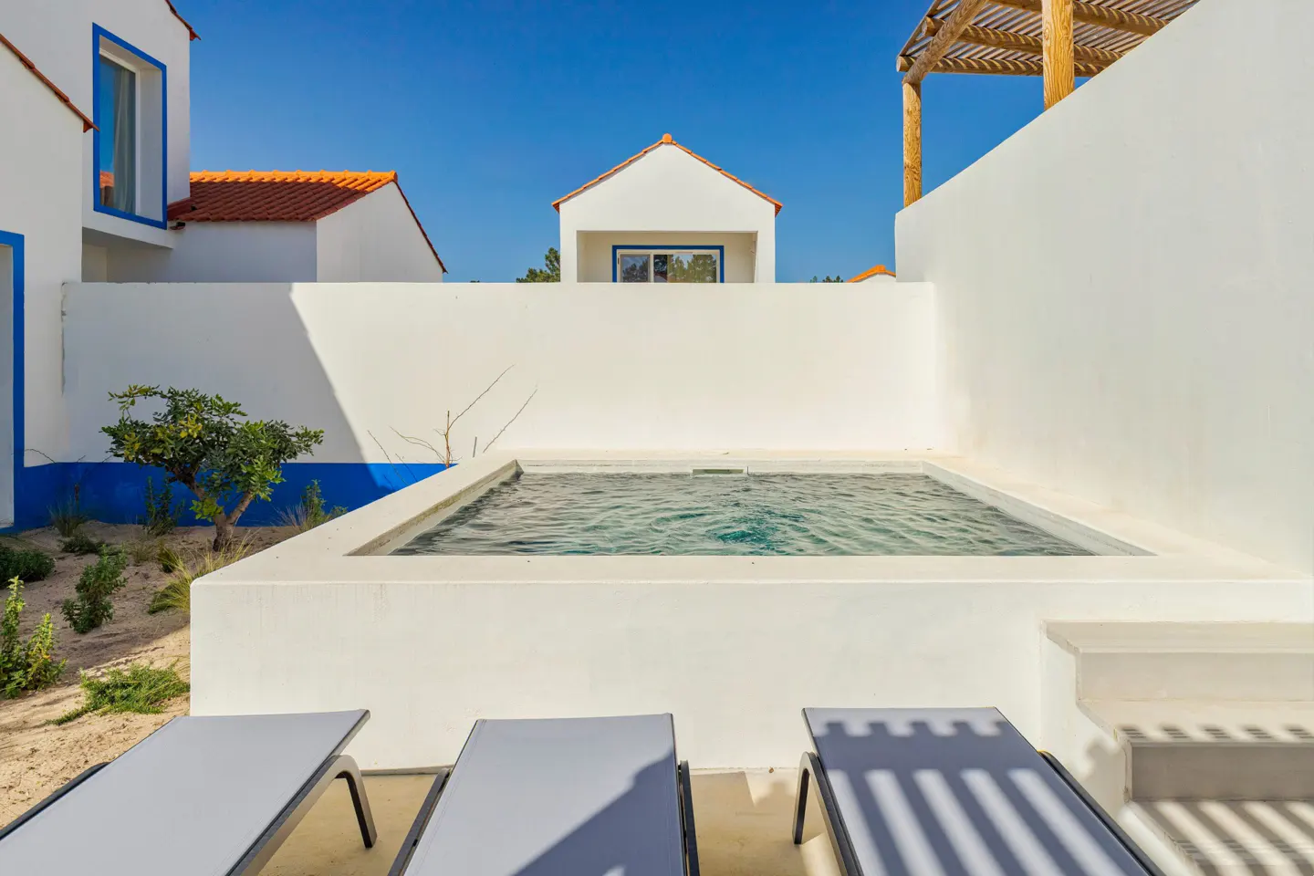 Outdoor view of a small pool surrounded by white walls, with lounge chairs in the foreground and white buildings with orange roofs in the background.