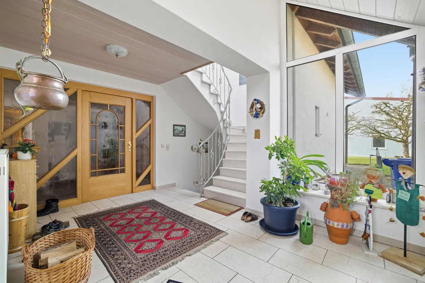 Bright foyer with white tile floor, wooden door, and winding staircase. A red patterned rug sits in the center. Plants and decorations adorn the window sill.