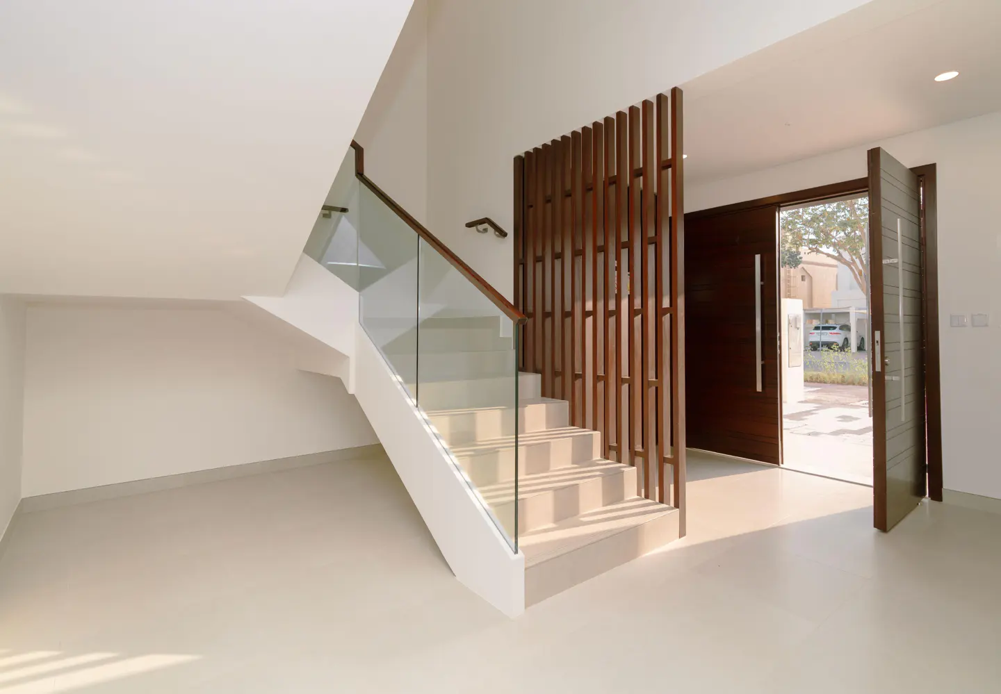 Bright foyer with white walls, beige tile floor, and a staircase with glass railing. Dark wood screen and open front door to the outside.