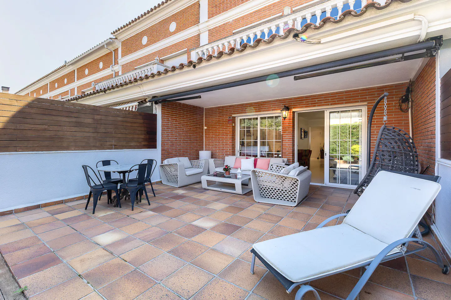 Outdoor patio with tile flooring, brick walls, and white trim. Lounge furniture, a dining table, and a hanging chair are arranged on the patio.