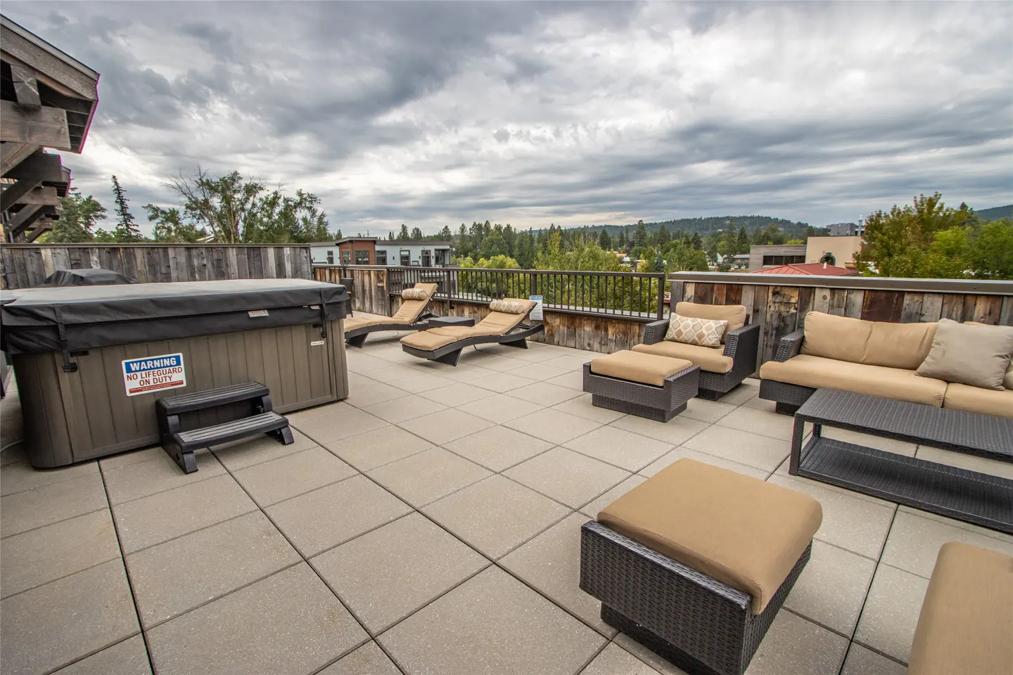 Rooftop patio with hot tub, lounge chairs, and seating area. City and trees in the background under a cloudy sky.