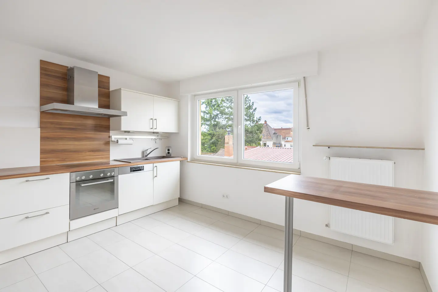 Bright kitchen with white cabinets, stainless steel appliances, and wood accents. A window overlooks a neighborhood with trees and houses. A small table stands in the foreground.