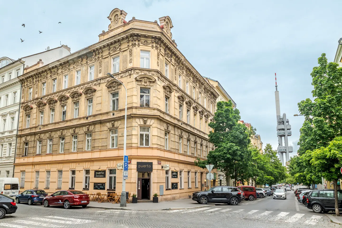 Street view of a tan corner building with "Puerto" restaurant, cars parked, and Žižkov Television Tower in the background.