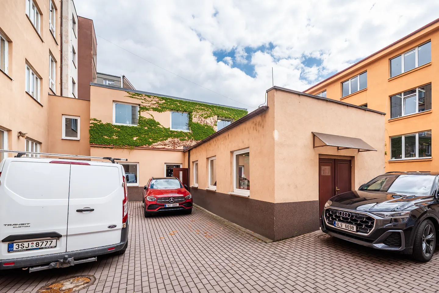 A courtyard with a white van, red Mercedes, and black Audi parked on a brick surface, surrounded by buildings.