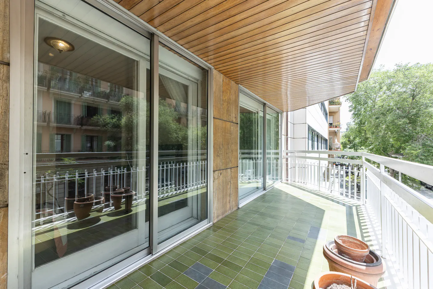 A balcony with green tile flooring, white railings, and sliding glass doors. Potted plants are visible.