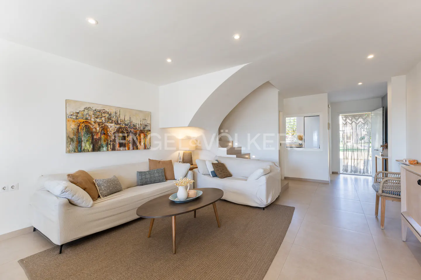 Bright living room with white sofas, brown rug, and a dark wood coffee table. A painting hangs above the sofa. Stairs lead to the upper level.